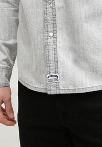 Chemise en jean gris clair à manches longues, boutons argentés et étiquette avec un texte bleu et blanc sur l'ourlet. Détails simples cousus.