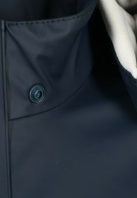 Navy blue vesken vedenkestävä takki kauluksella, jossa on nappikiinnitys. Takissa on sileä ulkopinta ja vaaleanvärinen, teksturoidulla vuorilla varustettu sisävuori.