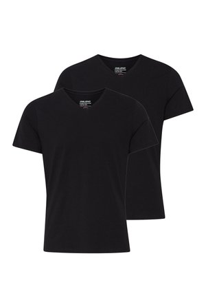 Homme debout face à l'avant, portant un t-shirt noir col V, un jean bleu et des baskets blanches sur un fond clair unicolore.