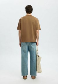 Hnědé oversized tričko, modré volné džíny, béžová kabelka a hnědé sandály. Hladký materiál, pohodlný střih, jednoduchý design.