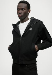 Čierna zipová bunda s kapucňou a šedým podšívkou; má malé logo na hrudi a elastické manžety. Kombinovaná s bielym tričkom pod tým.