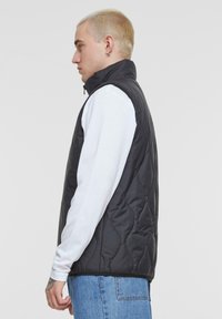 Schwarzige, gesteppte Weste mit hohem Kragen, Reißverschluss und Seitentaschen, getragen über einem weißen Langarmshirt und blauen Jeans.