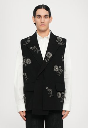 Sort vest med blomster sølvbroderi, struktureret design, to forlommer, båret over en hvid skjorte med spidse kraver.