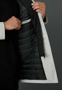 Chaqueta aislante con un exterior blanco y un forro acolchado negro. Presenta un bolsillo oculto con un ribete blanco en contraste y cremallera.