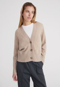 Cardigan beige realizzato in materiale morbido con scollo a V, orlo a coste e quattro bottoni in legno. Abbinato a pantaloni grigi su misura.