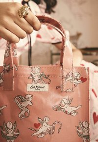 Rosa Tragetasche mit Cherubim- und Herzmustern, ausgestattet mit einer glatten Kunststoffoberfläche und doppelten Griffen. Das Etikett lautet "Cath Kidston London".