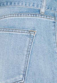 Vaaleansininen farkkukangas, jossa on näkyvä vinoneten kudonta ja takatasku, joka onommeltu kontrastivärisellä vaaleanruskealla langalla.