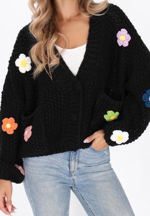 Femme portant un cardigan noir en maille avec des fleurs colorées en crochet, associé à un jean bleu clair et un haut blanc.