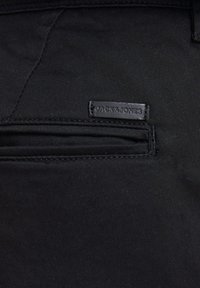 Pantalon en tissu noir avec une texture mate et élégante. Comprend une petite étiquette en cuir noir portant l'inscription "JACK & JONES" près de la poche zippée.