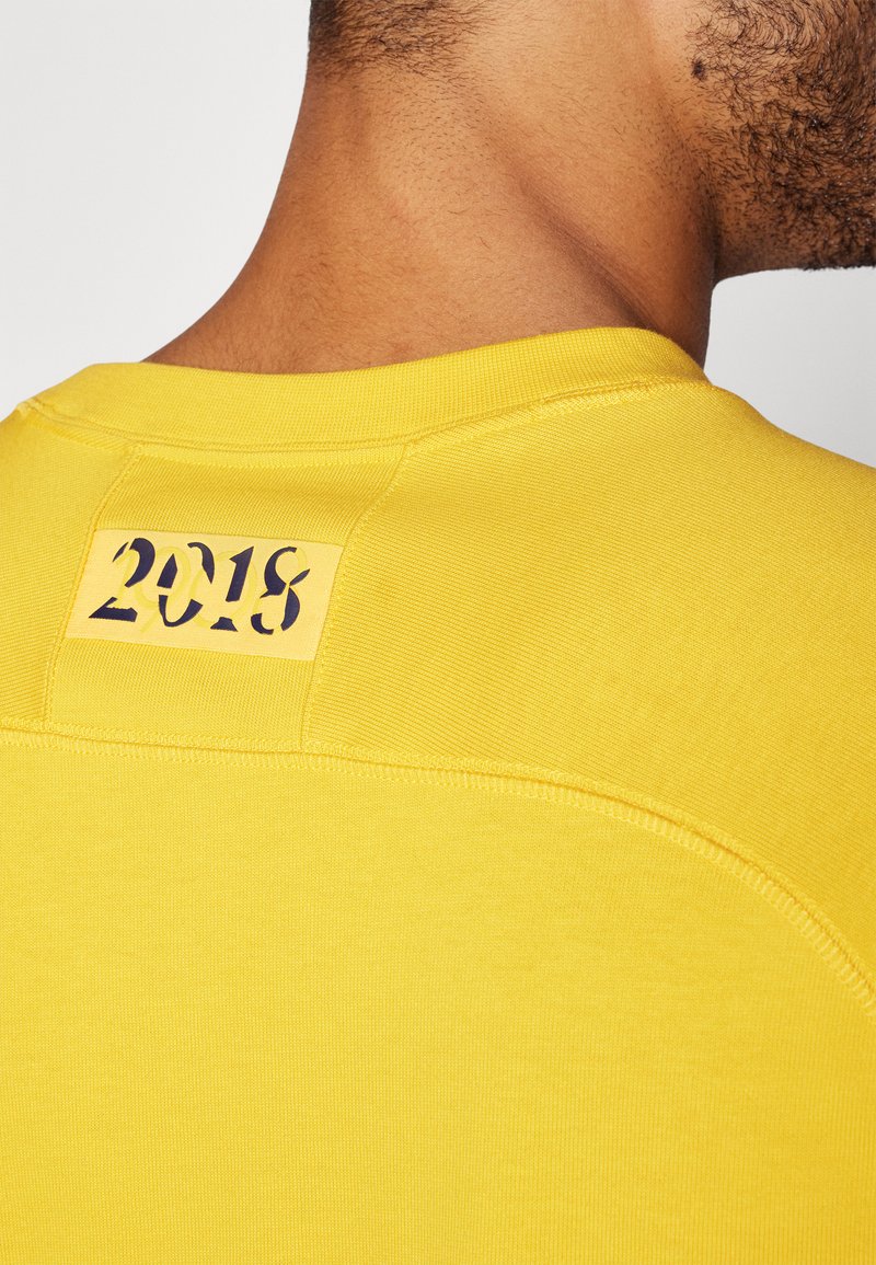 T-shirt giallo con scollatura rotonda. Presenta un'etichetta sul retro con la scritta "2018" in un colore scuro a contrasto. Texture liscia.