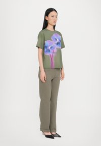 Olivengrünes T-Shirt mit kurzen Ärmeln, das mit einem lebhaften blauen und lila Blumenmuster verziert ist, kombiniert mit olivgrünen, figurbetonten Hosen und schwarzen Absätzen.