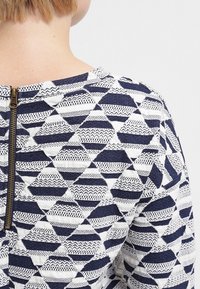 Haut à motifs bleu marine et blanc présentant des formes géométriques en losange, un tissu texturé et une fermeture éclair visible à l'arrière.