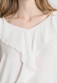 Blusa bianca con scollo a V e dettaglio frontale arricciato. Realizzata in tessuto leggero con una sottile trama. Le maniche corte non sono visibili.