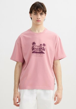 Joven con cabello castaño corto que lleva una camiseta rosa suelta con un gráfico de villa, palmeras y piscina en púrpura oscuro, combinada con pantalones blancos.