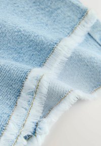 Közelkép világoskék farmer anyagról, dupla sor fehér foszlott varrással és látható aranyszínű varrással.