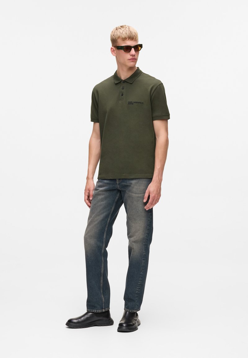 Olive green polo shirt with a collar, short sleeves, and embroidered logo. Paired with faded blue jeans and black footwear. Textured fabric.