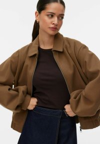 Blouson aviateur en polaire marron avec une fermeture éclair à l'avant, coupe décontractée et poignets côtelés, associé à un haut sombre et une jupe en denim.