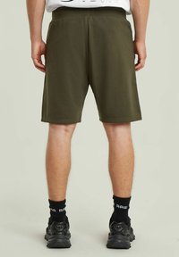 Pantaloni corti verde oliva realizzati in tessuto morbido, con vita elastica e una vestibilità rilassata, che terminano sopra il ginocchio. Sneakers nere e calze visibili.