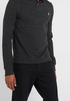 Mann trägt ein dunkelgraues langärmeliges Poloshirt mit einem kleinen weißen Logo auf der linken Brust und schwarze Hose, steht vor einem einfarbigen Hintergrund.