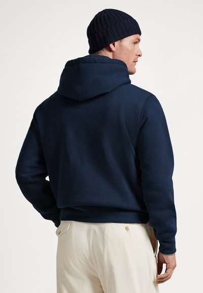 Mann iført en marineblå hettegenser og svart strikket lue, sett fra siden, kombinert med lyse beige kordfløyelbukser med knappede baklommer.