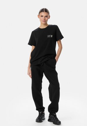 Naine seisab musta T-särgi, valge tekstiga ja mustade jooksupükstega, mustade spordijalanõudega, ühevärvilise valge tausta ees.