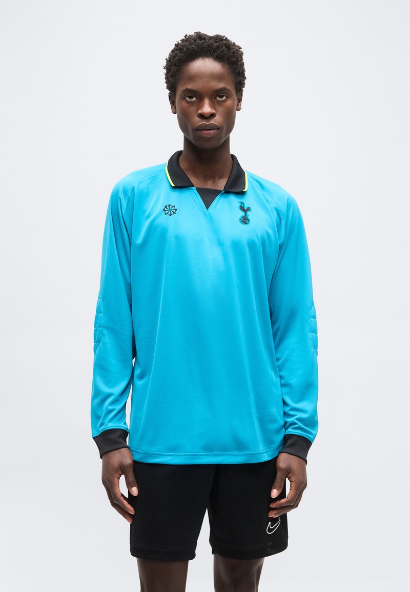 Νεαρός άνδρας φοράει αθλητική μπλούζα μακρυμάνικη ανοιχτού γαλάζιου με μαύρο γιακά και μαύρο σορτς, σε απλό λευκό φόντο.