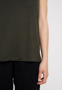 Top senza maniche di un verde scuro, realizzato in tessuto morbido, con orlo dritto e dettagli di cucitura minimi, abbinato a pantaloni neri testurizzati.