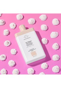 Eine Flasche mit aufgeschlagener Körperlotion auf einem rosa Hintergrund, umgeben von Klecksen weißer Creme, was die Produkttextur und das Design betont.