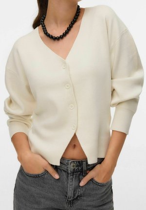 Frau trägt eine cremefarbene V-Ausschnitt-Strickjacke mit Knöpfen und Puffärmeln, eine schwarze Perlenkette und hoch sitzende, ausgewaschene schwarze Jeans, die Hände in den Taschen.