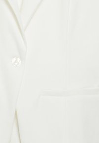 Valge bleiseri lähedalt vaade, kus on näha nööp, krae ja tikitud esitasku siledal kangal.