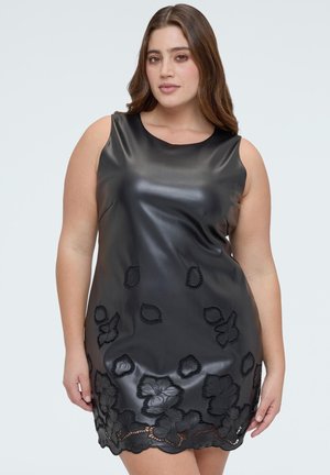 Frau trägt ärmelloses schwarzes Lederkleid mit floralen Spitzenausschnitten nahe dem Saum, steht vor einem schlichten hellen Hintergrund.