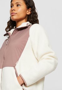 Veste en polaire bicolore avec un corps blanc crémeux et un panneau avant doux en mauve. Fermeture zippée, poches latérales et texture moelleuse.
