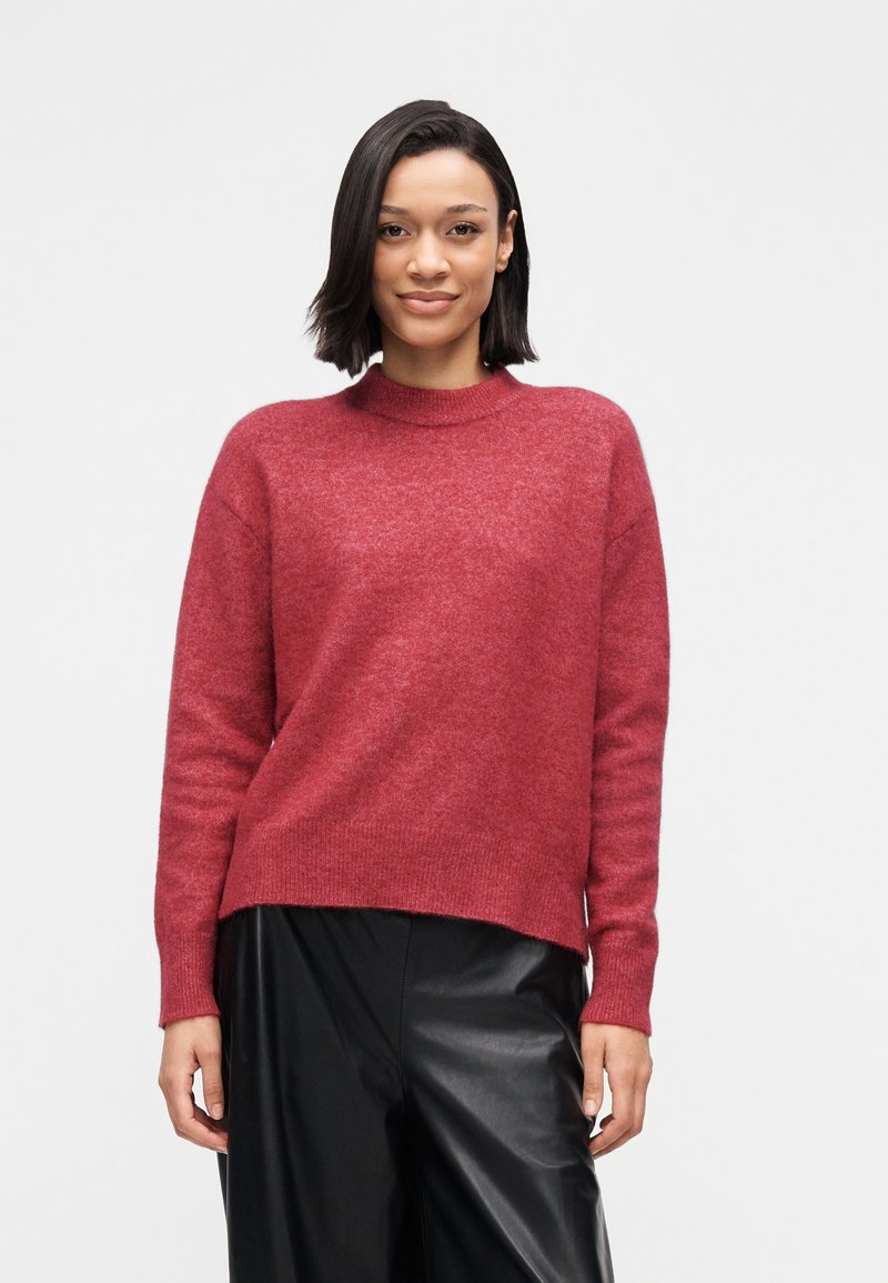 Pull en laine rouge avec un col montant, des épaules tombantes et une coupe décontractée. Associé à un pantalon noir en simili cuir.