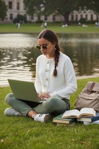 Young woman with sunglasses sitting cross-legged on grass by a lake, working on a laptop with books and a backpack nearby.