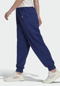 Marineblauwe trainingsbroek met elastische boorden, voorzien van zijzakken en een gouden logo-detail. Gedragen met witte sportschoenen.