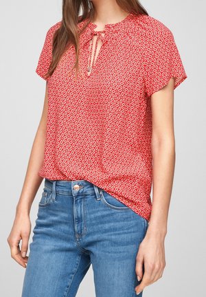 Mujer con blusa roja de manga corta con estampado y escote con abertura, y vaqueros azules de cintura alta sobre fondo liso.