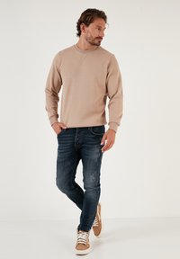 Felpa beige con scollo a girocollo, jeans strappati blu scuro e scarpe da ginnastica marroni con suola bianca. Design semplice e informale.