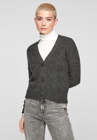 Cardigan en tricot gris avec un décolleté en V, présentant trois grands boutons noirs et une texture côtelée. Porté par-dessus un haut à col roulé blanc.