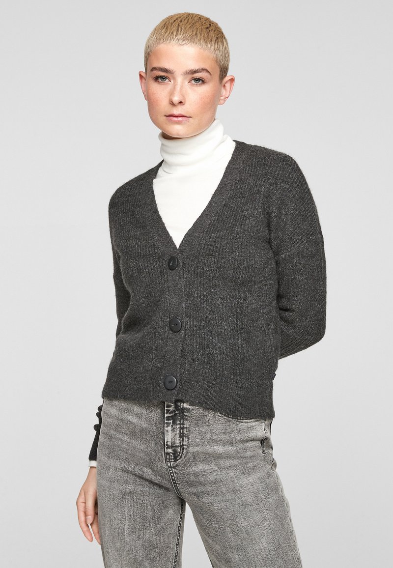 Cardigan en tricot gris avec un décolleté en V, présentant trois grands boutons noirs et une texture côtelée. Porté par-dessus un haut à col roulé blanc.