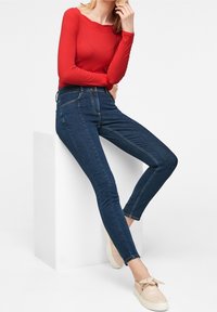 Czerwona bluzka z długim rękawem, ciemnoniebieskie rurki z wysokim stanem oraz jasne sneakersy z kokardką, na białym tle.