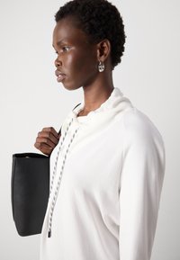 Witte hoodie met een gestructureerde afwerking en een gerimpelde kraag; heeft een zwarte koord met witte accenten. Model houdt een kleine zwarte tas vast.
