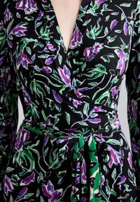 Květinové šaty s překřížením, které mají černý základ s fialovými a zelenými botanickými vzory, uvázané v pase s odpovídajícím páskem ze stejného materiálu.