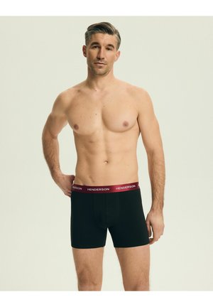Mężczyzna bez koszulki, o krótkich włosach, ubrany w czarne bokserki z czerwonym pasem z napisem "Henderson", stojący na jednolitym jasnym tle.