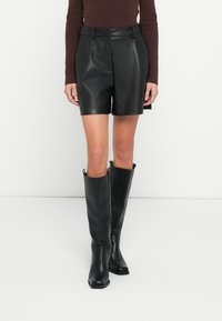 Zwarte leren shorts met een riem in de taille, voorzien van plooien en zijzakken, gecombineerd met zwarte leren kniehoge laarzen. Gladde textuur.