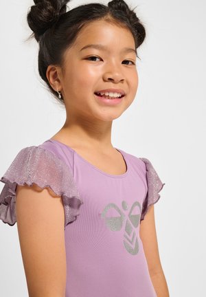 Młoda dziewczyna z dwoma kokami, ubrana w fioletową bluzkę z falbankami i błyszczącymi rękawami oraz srebrnym geometrycznym wzorem, lekko uśmiechająca się na bok.