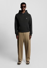 Sort hættejakke med en frontlomme, båret over beige, vide bukser. Solbriller og mønstrede sneakers fuldender outfittet.