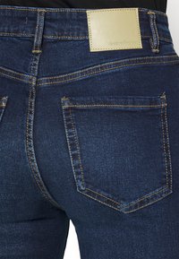 Jeans de denim azul escuro com costuras douradas contrastantes. Possui um retângulo de pele em tom de caramelo e dois bolsos traseiros com abas pontiagudas.