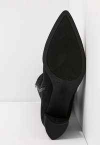 Escarpin noir à bout pointu avec semelle texturée, taille 37, placé debout contre des murs blancs d'angle.