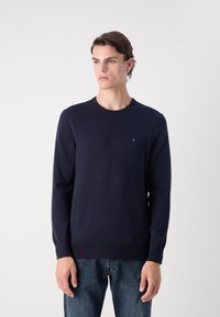 Marineblauer Strickpullover mit langen Ärmeln und Rundhalsausschnitt, versehen mit einem kleinen Logo auf der Brust. Der Stoff wirkt glatt und gleichmäßig.