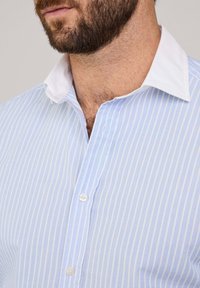 Camisa a rayas azul claro con cuello blanco, tapeta de botones y textura suave, que destaca por las líneas verticales y los botones redondeados.